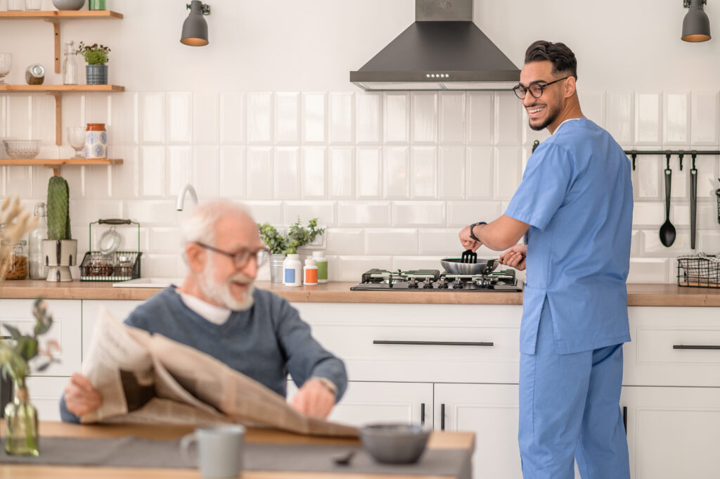 Homecare worker cooking at a stove while an older adult reads the newspaper nearby.